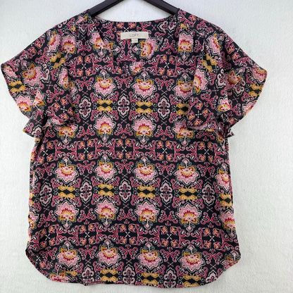 A short-sleeve blouse with a colorful paisley and floral pattern on a black background, hanging on a black hanger against a white textured wall.
