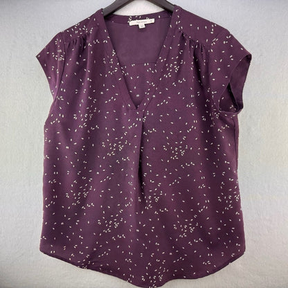 A purple short-sleeve blouse with a white dash pattern, hanging on a black hanger against a textured light gray wall.