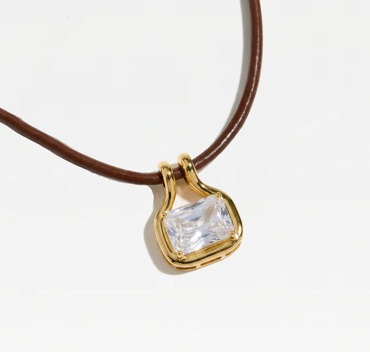 A close-up of a gold pendant with a rectangular clear gemstone, attached to a brown cord necklace, set against a plain white background.